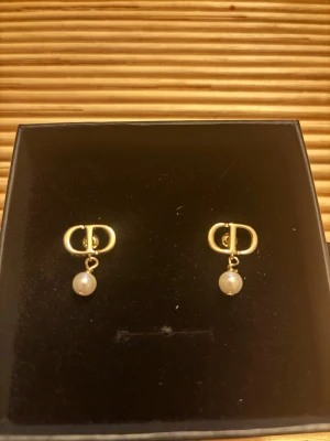 Petit Cd logo earring with goldfinish and resinDior - Snygga örhängen från Dior i guldfärg med klassisk CD-logga och hängande vita pärla (resin). Perfekta för dig som vill ha en elegant och trendig accessoar med exklusiv känsla. Stiftmodell som sitter bekvämt på örat. Vintage, minst 10 år. Har ej ngt kvitto som bevisar dess äkthet, men Vestiaire Collective (känd återsäljare av Dior och andra märken) godkänner de som äkta Dior. 