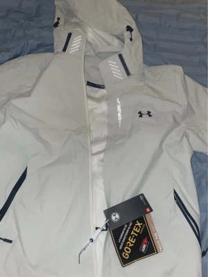 Vit GORE-TEX regnjacka Under Armour - Snygg vit regnjacka från Under Armour med GORE-TEX Paclite Plus-material som håller dig torr. Jackan har huva, dragkedja framtill, svarta detaljer och reflekterande tryck. Perfekt för regniga dagar och outdoor-äventyr.