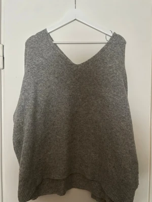 Grå stickad tröja från Pull&Bear - Mysig grå stickad tröja från Pull&Bear med v-ringning och avslappnad passform. Perfekt att slänga över en skjorta eller t-shirt för en chill lager-på-lager-look. Tillverkad i mjukt material som känns skönt mot huden.