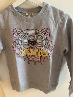 Grå sweatshirt med tiger från Kenzo - Säljer en grå sweatshirt från Kenzo med det ikoniska tigerhuvudet broderat framtill i svart, vitt, rosa och gult. Tröjan har rund halsringning, ribbade muddar och en avslappnad passform. Perfekt statement-plagg för dig som gillar streetstyle.