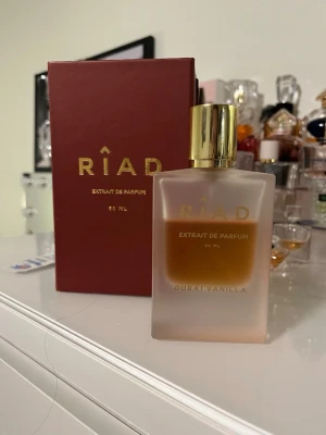 Riad Dubai Vanilla Extrait de Parfum - Riad Dubai Vanilla Extrait de Parfum är en lyxig parfym i en frostad glasflaska med guldfärgat lock. Doften är vaniljbaserad och flaskan rymmer 50 ml. Förpackningen är elegant vinröd med guldfärgade detaljer och text.