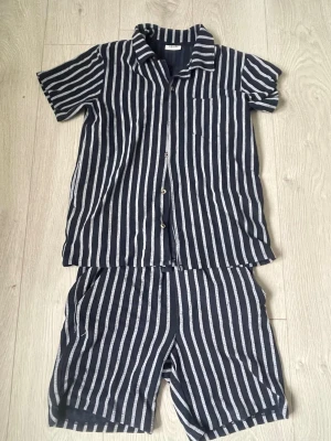 Randig pyjamas med shorts från Kappahl - Snyggt tvådelat somrigt set från Kappahl med kortärmad piké och shorts. Setet är marinblått med vita vertikala ränder och har knappar framtill på skjortan samt en bröstficka. Perfekt för chill dagar eller sommarnätter.