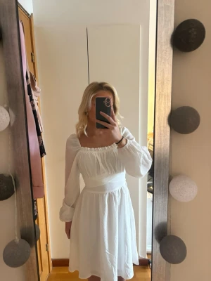 Vit klänning med puffärm och rosett - Supersöt vit klänning med långa puffärmar, markerad midja och en stor rosett i ryggen. Klänningen har en lätt rynkad överdel och är gjord i ett tunt, luftigt tyg. Perfekt till sommaren, studenter och högtider.