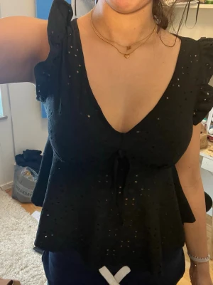 Svart blus med volangärm och knyt - Säljer en svart blus med v-ringning, volangärmar och dekorativt knyt under bysten. Blusen har broderade hål och är tillverkad i ett lätt tyg som ger en luftig känsla. Perfekt för dig som gillar feminina detaljer och en trendig look.