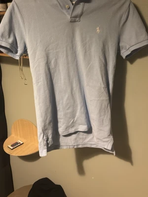 Ljusblå pikétröja från Ralph Lauren - Klassisk ljusblå pikétröja från Ralph Lauren med korta ärmar och vit broderad logga på bröstet. Tröjan har krage och knappar vid halsen. Tillverkad i mjuk bomull för en bekväm känsla.