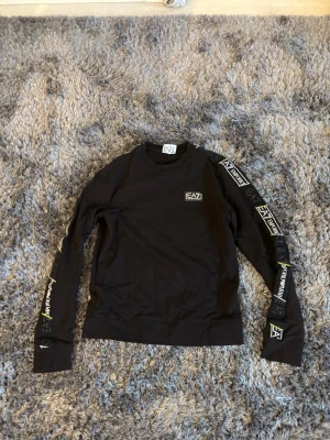 Svart långärmad tröja EA7 - Svart långärmad tröja från EA7 Emporio Armani med tryckta loggor längs båda ärmarna och en liten logga på bröstet. Tröjan har rund halsringning och är gjord i mjukt bomullsmaterial. Perfekt för en sportig och trendig look.