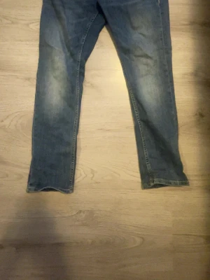 Blå raka jeans med slitningar - Säljer ett par blå jeans med raka ben och snygga slitningar på framsidan och baksidan. Jeansen har klassisk femficksdesign och en läderpatch bak i midjan. Perfekta för en avslappnad och trendig look.