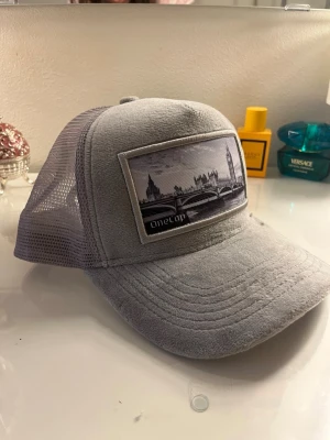 Ljusgrå keps LONDON - Säljer en ljusgrå keps från Onecap med mesh på sidorna och baksidan. Framtill finns en stor patch med ett svartvitt motiv av London och Big Ben. Kepsen har böjd skärm och justerbar snapback. Perfekt för dig som gillar streetstyle och coola detaljer.