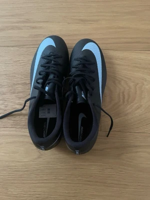 Nike Mercurial svarta fotbollsskor - Säljer ett par svarta Nike Mercurial fotbollsskor med vit swoosh och ljusblå dobbar. Skorna har snörning och är tillverkade i syntetmaterial för bra grepp och kontroll på planen. 