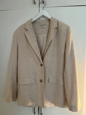 Beige linnekavaj - Stilren beige linne kavaj. Klassisk passform med två knappar framtill, fyra knappar vid ärmslut och tre fickor med lock. 