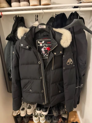 Svart dunjacka från Moose Knuckles - Svart dunjacka från Moose Knuckles med pälskant på huvan och flera fickor med dragkedja. Jackan har en cool logotyp på ärmen och ett grafiskt foder. Perfekt för kalla vinterdagar och har en modern, sportig look. Originalgaljen medföljer.