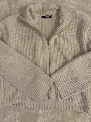 Beige stickad ziptröja från Bik Bok - Säljer en beige stickad ziptröja från Bik Bok med hög krage och grov ribbstickning. Tröjan har långa ärmar och dragkedja framtill, perfekt för lager på lager och en chill look.