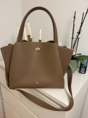 Brun handväska från Molly Tote oanvänd - Snygg brun handväska från Molly Tote i svagt strukturerat läder. Väskan har både handtag och en lång axelrem, samt guldfärgade detaljer och ett vridlås framtill. Helt oanvänd ❤️