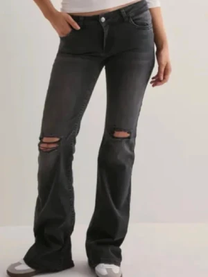 Svarta slitna bootcut jeans - Säljer ett par svarta bootcut jeans med slitningar och hål på båda knäna. De är lågmidjade och bootcut. De är använda ett fåtal gånger och har inga större skador förutom lite slitningar längst nere vid fötterna. Storlek 36 från Nelly.