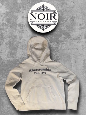 Vit hoodie från Abercrombie - Snygg vit hoodie från Abercrombie med trycket 'Abercrombie Est. 1892' på bröstet. Klassisk känguruficka framtill och justerbar huva med dragsko. Perfekt för en avslappnad och trendig look.