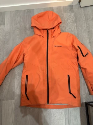 Skidjacka från Peak Performance - Snygg orange vindjacka från Peak Performance med huva och svarta dragkedjor. Jackan har tre framfickor och är  Perfekt för blåsiga dagar och outdoor-aktiviteter. Man kan diskutera om priset om du vill!!!