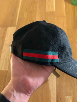 Svart keps från Gucci med röd/grön detalj - Snygg svart keps från Gucci med diskret GG-mönster i tyget och klassisk röd/grön rand på sidan. Justerbar metallspänne bak för perfekt passform. Perfekt accessoar för dig som vill ha en exklusiv och trendig look.