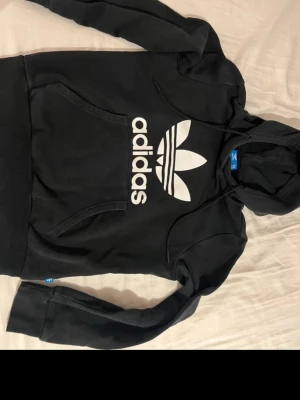 Svart Adidas hoodie med stor logga - Svart hoodie från Adidas med stor vit Trefoil-logga och text på bröstet. Klassisk känguruficka framtill och huva med dragsko. Tillverkad i mjuk bomullsblandning, perfekt för en avslappnad streetwear-look.