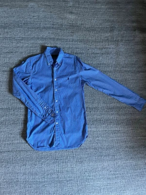 Blå randig skjorta från Ralph Lauren - Blå skjorta från Ralph Lauren med vita och mörkblå ränder. Slim fit-modell med klassisk krage och knappar framtill. Skick 9/10. Hör av dig vid minsta fundering! 