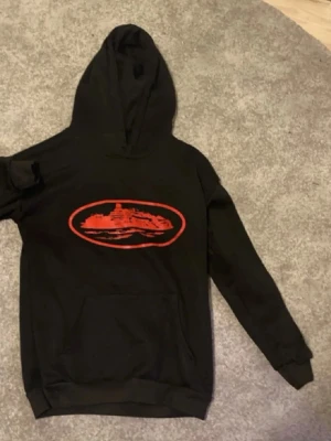 Cortiez Hoodie - Cool svart hoodie med ett stort rött tryck av ett skepp i oval ram på bröstet. Klassisk passform med huva och känguruficka framtill. Materialet ser ut att vara mjukt och bekvämt, perfekt för att chilla eller hänga med kompisar.