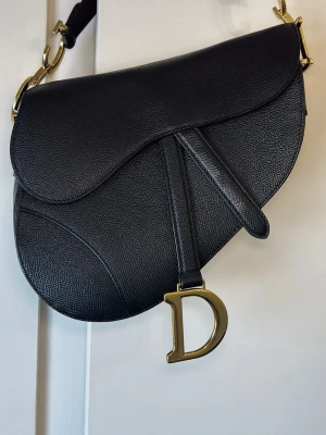 Saddle bag trendig väska - Saddle bag i svart kalvskinn, mått ca längd 25.5 höjd 20 djup 6.5cm. Så kallat "grained" läder vilket gör att eventuella märken bara är att torka bort. Använd fåtal gånger så i fint skick. 