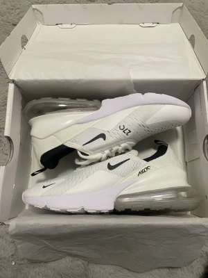 Nike Air Max 270 vita sneakers - Nike Air Max 270 sneakers i vitt med svarta detaljer och stor synlig Air-enhet i hälen. Ovandelen är i mesh för bra ventilation och har en modern, sportig look. Klassisk Nike Swoosh på sidan och AIR270-tryck på plösen och hälen. Perfekta för dig som gillar stilrena och bekväma sneakers.