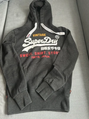Mörkgrå Superdry hoodie med tryck - Snygg mörkgrå hoodie från Superdry med stor vit och gul logga samt japansk text på bröstet. Hoodien har vita dragsnören, känguruficka och ribbade muddar. Perfekt för dig som gillar streetwear och vill ha en bekväm och stilren look. Aldrig använd.