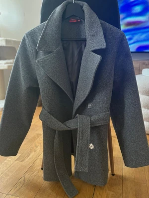 Kappa - Stilren mörkgrå kappa med knäppning framtill, två rymliga fickor och matchande midjeskärp. Använd fåtal gånger, väldigt fint skick!🩶 Storlek 158/164 (passar mig som är XS/S) 