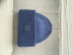 Mörkblå mössa från Acne Studios - Säljer en mörkblå ribbstickad mössa från Acne Studios med uppvikt kant och det klassiska ansiktslogotypen framtill. Mössan är gjord i tjockt, mjukt material och har en modern, avslappnad passform. Perfekt för kalla dagar och stilrena outfits.