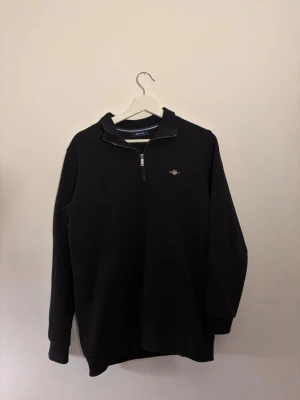 Svart half-zip tröja från GANT - Snygg svart tröja från GANT med half-zip dragkedja och liten broderad logga på bröstet. Klassisk rak passform och ribbade muddar vid ärmslut och nederkant. Perfekt till jeans eller chinos för en clean look.