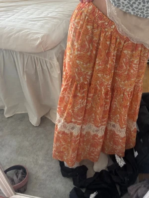 Orange långkjol med spetsdetaljer - Bohemisk långkjol i orange med paisleyinspirerat mönster och breda vita spetsdetaljer. Kjolen har en luftig passform och resår i midjan, perfekt för sommarens alla dagar.