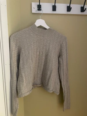 Beige kabelstickad tröja - Snygg beige kabelstickad tröja med högre ribbad hals och långa ärmar. Tröjan har ett klassiskt mönster och ribbade muddar vid ärmslut. Perfekt för lager på lager och enkel att matcha till olika outfits.