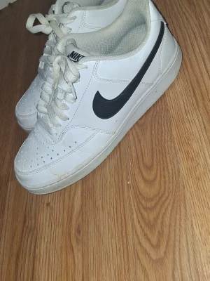 Vita Nike sneakers med svart swoosh - Säljer ett par klassiska vita Nike sneakers med svart swoosh på sidan. Skorna har lågt skaft, vita snören och ventilerande hål på tån. Tillverkade i syntetmaterial och har en tidlös streetstil som passar till det mesta.