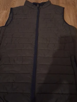 Mörkgrön dunväst med dragkedja - Stilren mörkgrön dunväst med horisontella sömmar och hel dragkedja framtill. Västen har en hög krage och är ärmlös, perfekt för lager-på-lager. Materialet är quiltat och ger en sportig look. Diskret logga på bröstet.