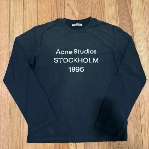 Svart långärmad Acne Studios t-shirt - Säljer en svart långärmad t-shirt från Acne Studios med vit text 'Acne Studios STOCKHOLM 1996' på bröstet. Klassisk rund hals och normal passform. Perfekt för dig som gillar stilrena och ikoniska plagg.