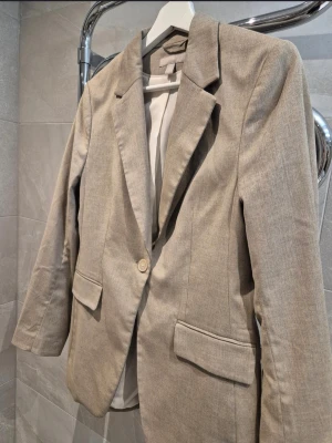 Beige kavaj med klassisk passform - Stilren beige kavaj med en knapp framtill och två fickor med lock. Kavajen har klassisk krage och är helfodrad. Perfekt för en snygg och enkel look. Materialet ser ut att vara en blandning av bomull och polyester.