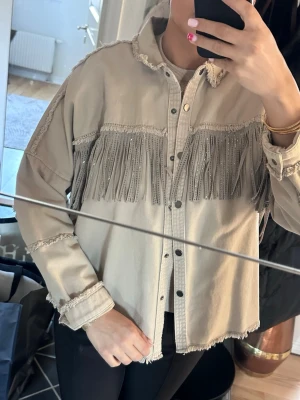 Beige jacka med fransar - Trendig beige skjorta med fransar över bröstet och armarna. Skjortan har knappar framtill och råa kanter vid krage, ärmslut och nederkant. Perfekt för dig som gillar western-inspirerad stil och vill sticka ut.