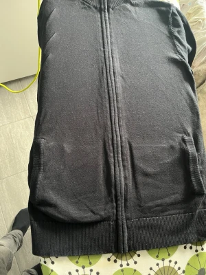 Svart stickad väst med dragkedja - Säljer en svart stickad väst med hel dragkedja framtill och ribbade muddar. Västen har fickor på sidorna och en enkel, stilren design som passar till många olika outfits.