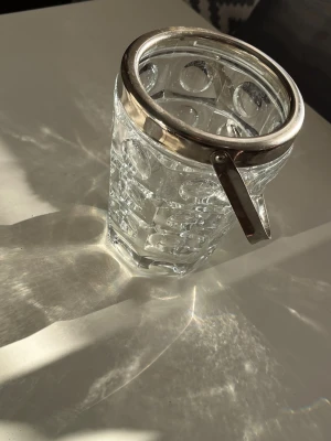 Glasburk med metallhandtag - Säljer en genomskinlig glasburk med dekorativt mönster och ett robust metallhandtag. Burken har en rund öppning och är perfekt för förvaring eller dekoration. Metallringen upptill ger en snygg kontrast till glaset.