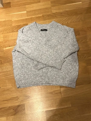 Ljusgrå stickad tröja FB Sister - Mysig ljusgrå stickad tröja från FB Sister med v-ringning och ribbade muddar vid ärmslut och nederkant. Perfekt för lager på lager och enkel att matcha med olika outfits. Tillverkad i mjukt material för extra komfort.