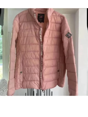 Rosa pufferjacka från Polo Club - Snygg rosa pufferjacka från Polo Club med hög krage och quiltad design. Jackan har dragkedja framtill, två sidofickor med tryckknappar och ett Polo Club-märke på ärmen. Perfekt för kalla dagar och ger en trendig look.