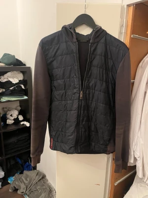Svart Prada cardigan  - Säljer min Prada cardigan. Jackan har huva och hel dragkedja framtill. Materialet är en mix av vadderad polyester på kroppen och bomull på ärmar och rygg. Perfekt för dig som gillar streetwear och vill ha något bekvämt och stilrent.