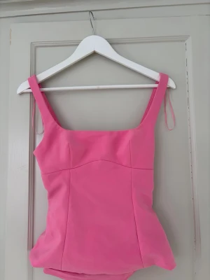 Rosa linnetopp med rosett i ryggen - Supersöt rosa linnetopp med breda axelband och en stor rosett i ryggen som ger en unik look. Toppen har en öppen rygg och är figurnära i modellen. Perfekt för dig som vill sticka ut lite extra i sommar.