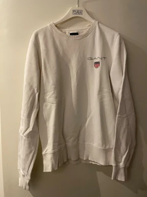 Vit sweatshirt från GANT - Klassisk vit sweatshirt från GANT med rund halsringning och broderad logga samt sköld på bröstet. Tröjan har ribbade muddar vid ärmslut och nederkant. Perfekt för en clean och stilren look.