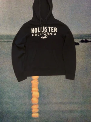 Marinblå  hoodie från Hollister - Svart hoodie från Hollister med stor vit logga och texten 'Hollister California' på bröstet. Klassisk känguruficka framtill och justerbar huva med snören. Mjuk insida och ribbade muddar vid ärmslut och nederkant. Hör av er om ni har några frågor.
