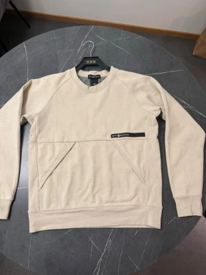 Beige sweatshirt från Sail Racing - Stilren beige sweatshirt från Sail Racing med svart logotypdetalj på bröstet. Tröjan har rund hals, långa ärmar och en unik fickdesign framtill. Perfekt för en avslappnad och trendig look.
