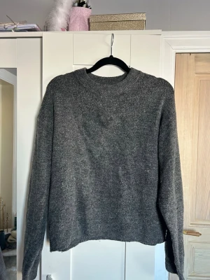 Mörkgrå stickad tröja från H&M - Mysig mörkgrå stickad tröja från H&M med rund halsringning och långa ärmar. Tröjan har en enkel, rak passform och ribbstickade muddar vid halsen. Perfekt för lager på lager under kyliga dagar.