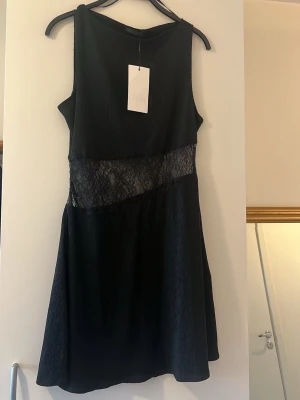 Svart klänning med spetsdetalj från Zara  - Snygg svart kortklänning från Zara med transparent spetsdetalj i midjan. Ärmlös modell med rund halsringning och lätt utställd kjol. Perfekt för dig som gillar stilrena plagg med en twist. Passar even till storlek M