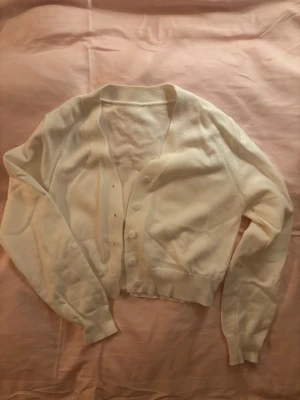 Vit cropped kofta med knappar - Säljer en vit cropped kofta med långa ärmar och ribbade muddar. Koftan har en klassisk V-ringning och stängs med vita knappar framtill. Perfekt att slänga över en topp för en enkel och clean look. Storlek S men känns som XS