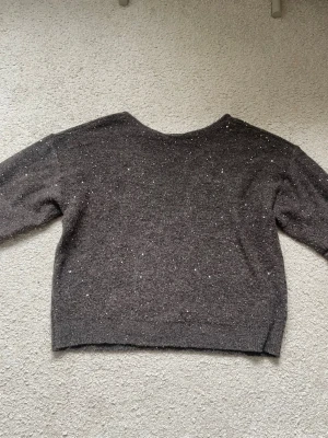 Mörkgrå stickad tröja med glitter - En mörkgrå stickad tröja med diskreta glitterdetaljer över hela plagget. Tröjan har en rund halsringning och långa ärmar, samt ribbstickad nederkant. Perfekt för dig som vill ha något enkelt men ändå lite extra.
